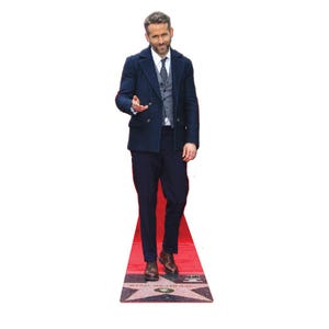 May include: A cardboard cutout of a man in a navy blue suit, standing on a red carpet. The cutout is on a white background. The man is wearing a navy blue coat, a gray vest, a white shirt, and a tie. The red carpet has a star with the name "RYAN REYNOLDS" on it.