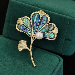 Op de afbeelding: Een goudkleurige metalen broche in de vorm van een ginkgoblad, voorzien van iriserende abalone schelpinleg in blauwe en groene tinten. Het ontwerp bevat een enkele witte parel en een dunne, met draad omwikkelde steel.