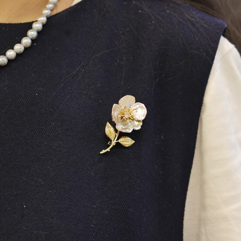 May include: A floral brooch featuring a pearlescent flower with a gold-colored stem and leaves. The flower has a cluster of small, sparkling accents at its center. The brooch is pinned to a dark navy blue garment.