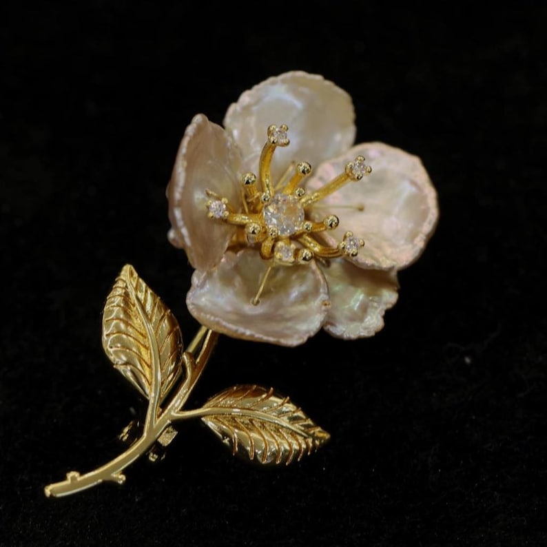 May include: A gold-toned flower brooch with a pearl-like petal design and a central clear gemstone. The brooch features a stem and leaves, all in a gold finish, set against a black background.