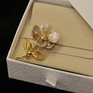 May include: A gold-toned floral brooch with pearl-like petals and a central diamond-like accent. The brooch features a detailed stem and leaves, presented in a white box with a beige interior, suggesting a jewelry item.