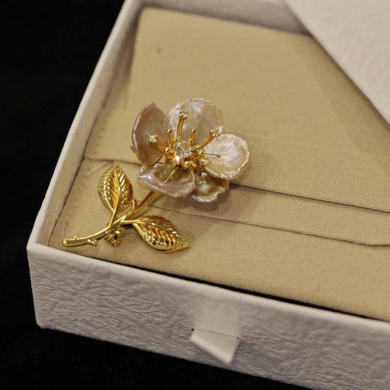 May include: A gold-toned floral brooch with pearl-like petals and a central diamond-like accent. The brooch features a detailed stem and leaves, presented in a white box with a beige interior, suggesting a jewelry item.