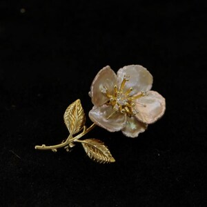 May include: A gold-toned flower brooch with a pearl-like center and diamond accents. The brooch features a detailed stem and leaves, set against a black background. This floral accessory is suitable for various occasions.