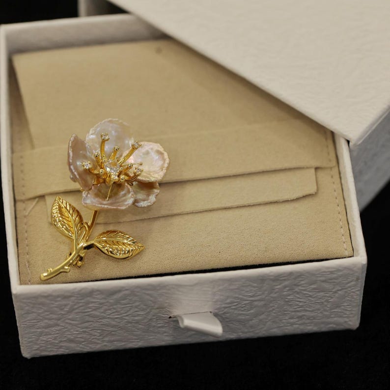 May include: A gold-toned flower brooch with pearl petals and diamond accents, resting in a white textured gift box with a beige interior. The brooch features a stem and leaves, adding to its floral design.