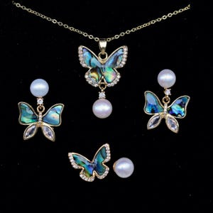 Puede incluir: Un conjunto de joyas doradas con colgantes y pendientes de mariposa. Las mariposas tienen alas iridiscentes azules y verdes, acentuadas con cristales transparentes. El conjunto incluye un collar con colgante y un par de pendientes, además de un par de pendientes de mariposa.