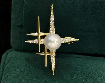 May include: A gold-toned brooch featuring a central white pearl surrounded by star-shaped accents. The design includes two smooth, polished gold stars and a larger, elongated star encrusted with small, clear, sparkling crystals.