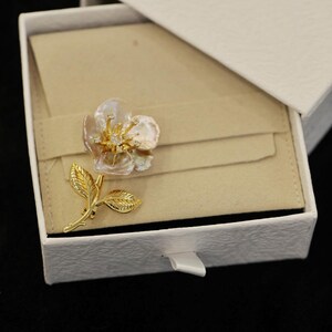 May include: A gold-toned floral brooch with a pearl-like center and small diamond accents, resting on a beige fabric inside a white textured box. The brooch features a detailed leaf and stem design.