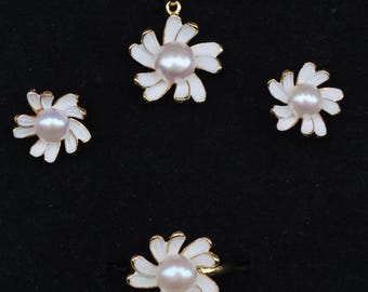 May include: A four-piece jewelry set featuring a daisy design. The set includes a pendant necklace, a pair of earrings, and a ring. Each piece has white enamel petals, gold-colored accents, and a central pearl. The jewelry is displayed on a black surface.