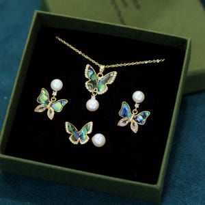 Könnte beinhalten: Ein Schmuckset mit Schmetterlingsmotiv, präsentiert in einer grünen Schachtel. Das Set umfasst eine Halskette, Ohrringe und einen Ring, alle aus goldfarbenem Metall und mit schillernden Schmetterlingsflügeln. Jedes Stück hat einen Perlenakzent.