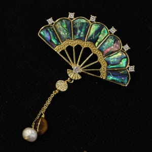 May include: A gold-coloured fan-shaped brooch with iridescent abalone shell segments. The brooch features a delicate filigree design, small clear crystals, and a dangling chain with a pearl and a brown stone. The background is black.