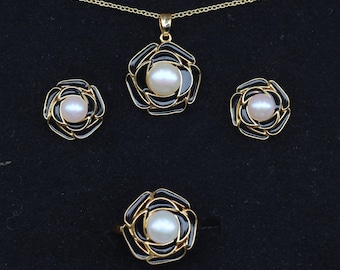 May include: A jewelry set featuring a pendant necklace, earrings, and a ring. Each piece has a gold-tone floral design with black enamel and a central pearl. The necklace chain and ring band are also gold-toned.