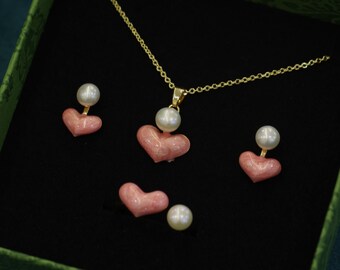 Puede incluir: Un conjunto de collar y pendientes dorados con diseño de corazón y perlas. El conjunto incluye un collar con colgante de corazón y un par de pendientes en forma de corazón. Los pendientes y el colgante son rosas con un acento de perla. Presentado en una caja verde.