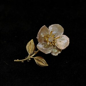 May include: A gold-toned floral brooch with a pearl-like flower and detailed leaves. The flower's center features a sparkling gem, and the petals have a textured appearance. The brooch is set against a black background.