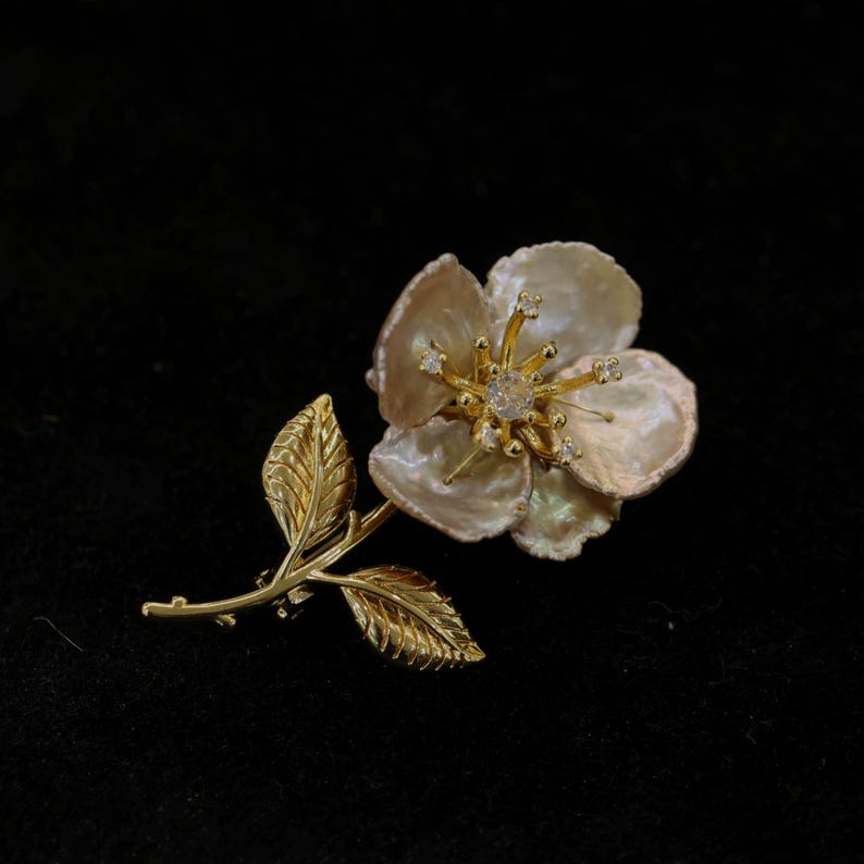 May include: A gold-toned floral brooch with a pearl-like flower and detailed leaves. The flower's center features a sparkling gem, and the petals have a textured appearance. The brooch is set against a black background.