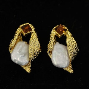 May include: A pair of gold-toned earrings with a textured leaf design. Each earring features a white, irregularly shaped pearl and a square, amber-colored gemstone at the top. The earrings are displayed against a black background.