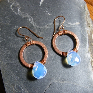 May include: Copper hoop earrings with a hammered texture, wrapped with copper wire. Each earring features a teardrop-shaped, faceted, opalescent blue gemstone. The earrings are displayed on a dark gray, textured surface.