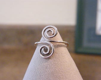 May include: A silver-colored ring with a double spiral design. The ring is displayed on a light-colored surface, showcasing its intricate, openwork pattern. The spirals are symmetrical and create a unique, artistic look.