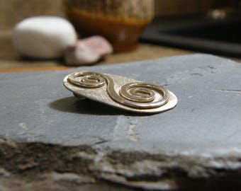 May include: A silver-toned metal brooch featuring an elongated oval shape with a textured surface and two raised, polished spiral designs connected in the center. The piece has a metallic sheen and a pin mechanism visible on the underside.