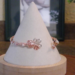 Könnte beinhalten: Handgefertigtes Armreif-Armband aus verflochtenem Kupfer- und silberfarbenem Draht. Das Armband hat dekorative Blumenakzente in Kupfer und Silber. Präsentiert auf einem weißen, kegelförmigen Schmuckständer.