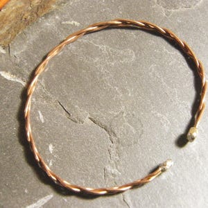 May include: A copper-coloured, open-ended bracelet with a twisted rope design. The bracelet is made of thin, intertwined strands of metal, creating a textured surface. The ends of the bracelet have small, rounded caps.