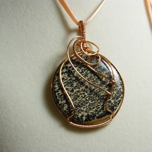 May include: A round pendant with a speckled black and white stone, wrapped in copper wire. The pendant hangs from a peach-colored ribbon. The wirework has an intricate design.