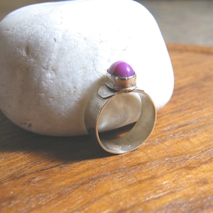 May include: A gold-toned ring with a large, round, purple gemstone set in a silver bezel. The ring is displayed on a white stone against a wooden surface. The ring's band is wide and smooth.