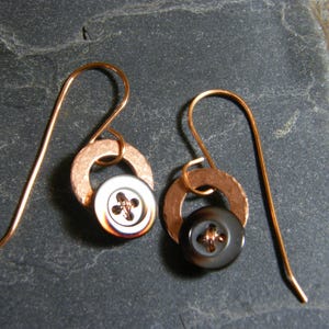 Puede incluir: Un par de pendientes hechos a mano con ganchos de cobre, anillos de cobre martillado y botones reutilizados. Un pendiente tiene un botón blanco con un diseño de cruz de cobre, el otro un botón gris oscuro. Los pendientes se muestran sobre una superficie gris oscura.