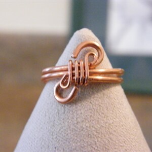 May include: A handcrafted copper wire ring with a unique design. The ring features a double band and decorative swirls and coils. The copper has a warm, reddish-brown hue. The ring is displayed on a light gray surface.