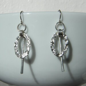 May include: A pair of silver-toned drop earrings featuring a textured, hammered oval hoop suspended from a circular ring, with a straight, smooth vertical bar extending downwards through the centre of each oval, attached to a standard hook wire.