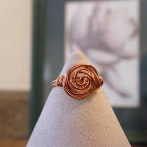 May include: A copper-colored wire ring with a spiral rose design. The ring is displayed on a light gray cone-shaped stand. The ring's design features a central spiral surrounded by coiled wire, creating a three-dimensional floral effect.