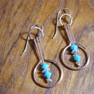 May include: A pair of handmade earrings featuring a copper-colored metal design with a teardrop shape. Each earring has a silver-colored hook and a cluster of turquoise-colored beads and silver-colored beads. The earrings are displayed on a wooden surface.