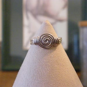 Könnte beinhalten: Ein silberfarbener Drahtring mit einem Spiraldesign in der Mitte. Der Ring wird auf einem beigefarbenen, kegelförmigen Ständer präsentiert. Die Spirale ist der Mittelpunkt, mit mehreren Drahtwicklungen, die das Band bilden.