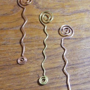 May include: Three handmade wire hair accessories with a spiral design. The wires are in copper, gold, and silver colors. Each accessory has a wavy stem and a spiral at the top and bottom. The background is a wooden surface.