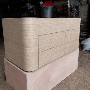 May include: A light beige dresser with a smooth, curved left side and six rectangular drawers arranged in two vertical columns. The dresser is constructed from a textured material with visible horizontal grain and rests on a plain wooden base.
