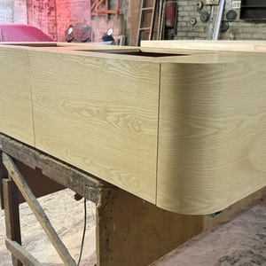 May include: A light-colored wooden cabinet with a curved edge. The cabinet is rectangular with three doors and is sitting on a wooden work table. The wood grain is visible. The cabinet is in a workshop setting.