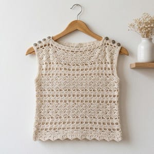 May include: A sleeveless cream-colored crochet top featuring an intricate openwork pattern with horizontal bands of shell-like stitches. The garment has a round neckline and is accented with three small metallic buttons on each shoulder seam.
