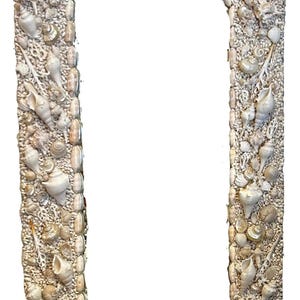 May include: An arched mirror frame, intricately decorated with a mosaic of various seashells. The frame is a light beige color, with a variety of shell shapes and sizes. The overall design evokes a coastal or nautical theme.