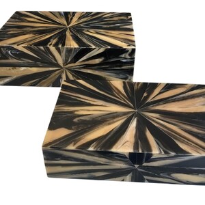 May include: Two rectangular decorative boxes with a striking starburst pattern. The boxes feature a combination of black and tan colors, creating a visually appealing contrast. The design is symmetrical, with the pattern radiating from the center of each box.
