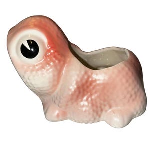 May include: A pink ceramic planter shaped like a creature with a large eye and a small opening at the top. The planter has a textured surface and a white interior. The eye is black and white.