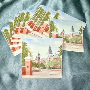 May include: A stack of square art prints featuring a watercolor illustration of a brick campus building with a tall spire and a prominent brick gate pillar in the foreground. The scene includes green trees, a paved path, and a signature at the bottom.