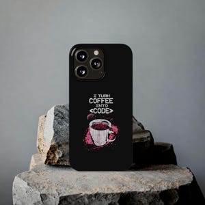 Peut inclure: Coque de téléphone noire avec un motif de tasse à café et le texte "I TURN COFFEE INTO CODE". Le motif de la tasse présente des accents rouges et blancs. La coque est présentée sur une surface en pierre grise.