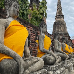 Puede incluir: Una fila de estatuas de Buda sentadas con túnicas amarillas, sobre un fondo de antiguas estructuras de ladrillo y altas agujas puntiagudas. Las estatuas son grises y el cielo está nublado.