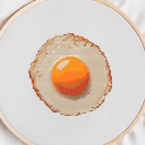 Op de afbeelding: Een witte borduurring met een kruissteekontwerp van een gebakken ei. De dooier is fel oranje.