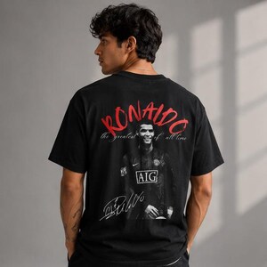 Puede incluir: Camiseta negra con un retrato de Cristiano Ronaldo y la palabra "RONALDO" en letras rojas. La camiseta también incluye la frase "the greatest of all time" y una firma impresa.