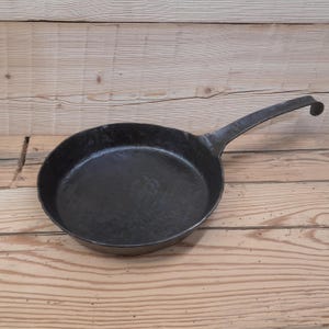 May include: A black, round, cast iron skillet with a long, curved handle. The pan is sitting on a wooden surface. The skillet is designed for cooking and has a hammered texture.
