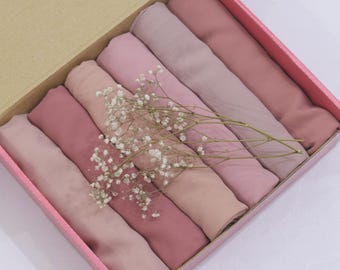 May include: A box of rolled fabric in various shades of pink and mauve. The fabric is arranged neatly inside a pink box, with a sprig of white flowers laid across the top. The box has a light brown lid.