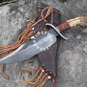 May include: A handmade knife with a brown leather sheath and a bone handle. The knife has a long, curved blade and a leather strap with beads and fringe.