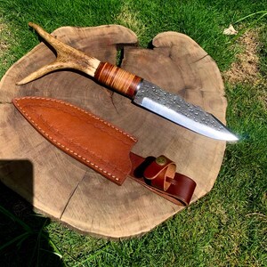 May include: A handcrafted hunting knife with a textured blade and a handle made of wood and antler. The knife is displayed with a brown leather sheath on a wooden surface, showcasing the craftsmanship and design.