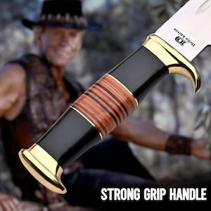 May include: Close-up of a knife with a polished steel blade and a handle featuring black, gold, and brown wood accents. The handle is inscribed with "STRONG GRIP HANDLE". The knife is from Delta Knives.