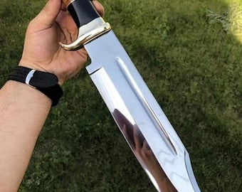 May include: A large fixed-blade knife with a polished silver-tone steel blade featuring a central fuller. The handle is constructed from alternating sections of black material and brown wood, accented by a gold-tone metal guard and pommel.